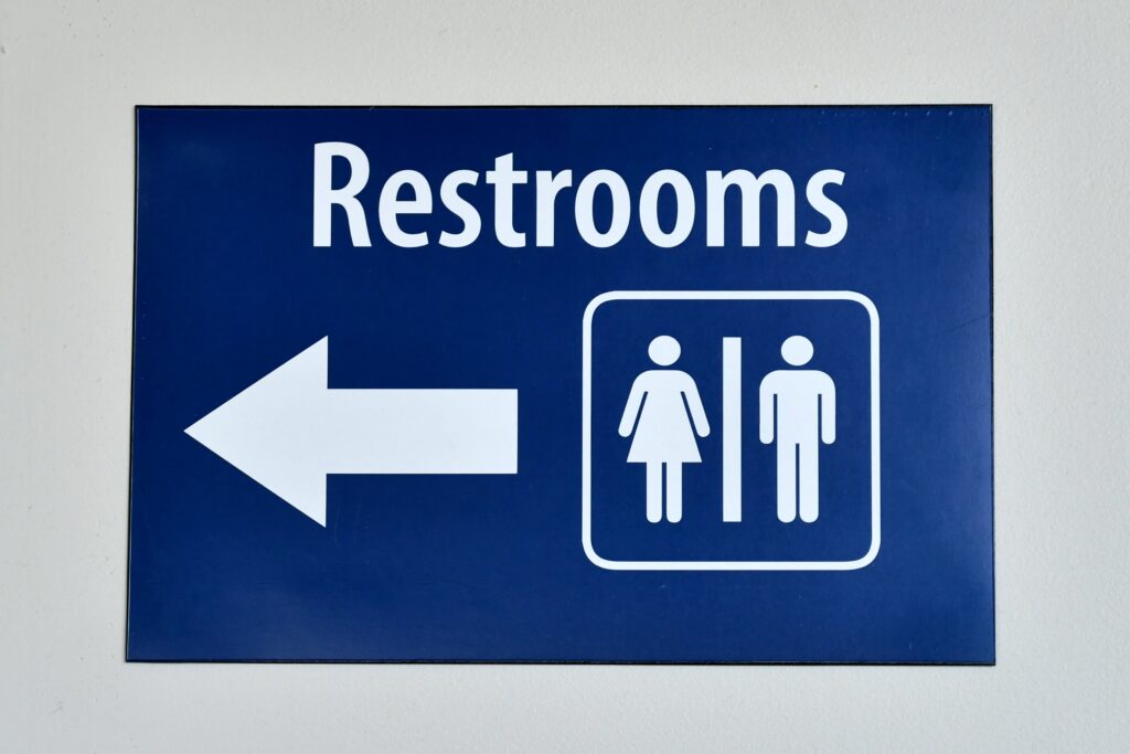 Blue restroom sign with white text indicating restrooms to the left, featuring icons of a woman and a man. Designed for easy access, it ensures everyone, including those experiencing incontinence, knows exactly where to go.