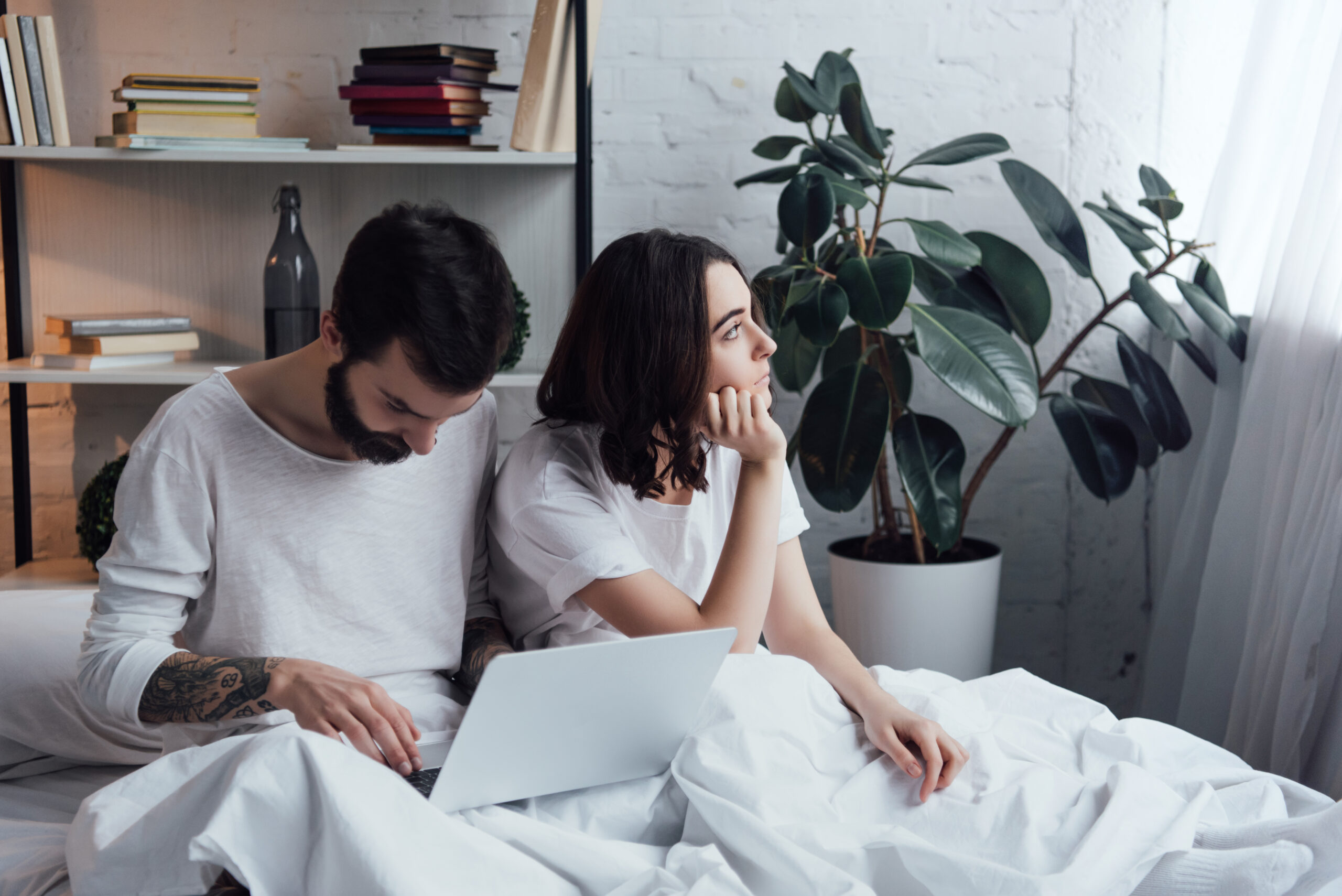 A man uses a laptop while sitting on a bed, possibly researching Erectile Dysfunction Treatment in Orlando, FL. The woman next to him looks out the window. They are surrounded by white bedding, a potted plant, and a bookshelf with stacked books.