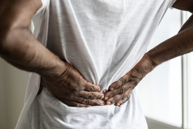 A person in a white shirt is holding their lower back with both hands, indicating back pain or discomfort, potentially seeking prostate cancer treatment in Orlando, FL.