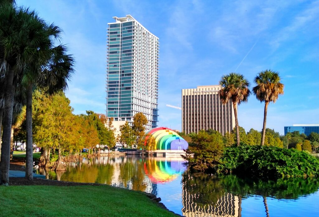 A cityscape featuring a tall modern building, a rainbow-colored bandshell, and palm trees. A lake with clear reflections of the buildings and trees is in the foreground.