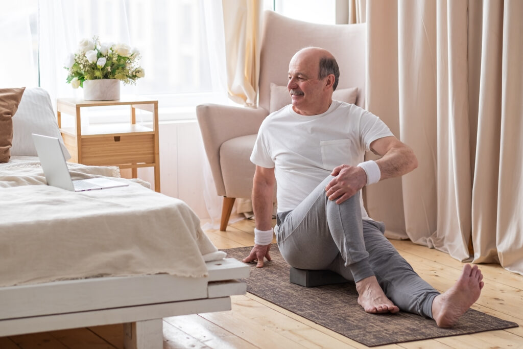 An older man sits on a yoga mat in a softly lit bedroom, stretching his legs while looking at a laptop. Dressed in comfortable workout clothes, he focuses on maintaining his health, perhaps with guidance from an FL Men's Health Clinic in Orlando. A bouquet of flowers adorns the bedside table.