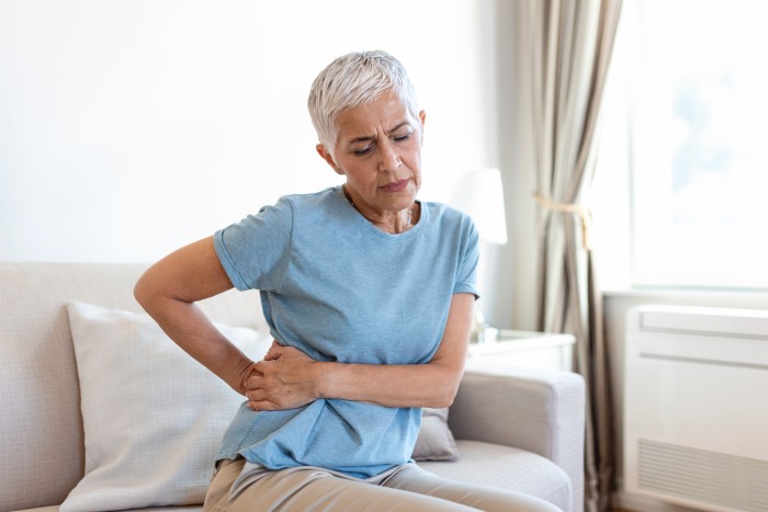 An older woman sits on the couch, pressing her lower back with a pained expression. She wears a light blue shirt, perhaps managing pelvic pain that's all too familiar in her routine.