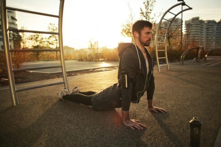 Man wearing headphones and athletic clothing doing a cobra stretch on an outdoor exercise area at sunrise, with urban buildings in the background—promoting wellness alongside Incontinence Treatment in Orlando: Effective Options for Relief.