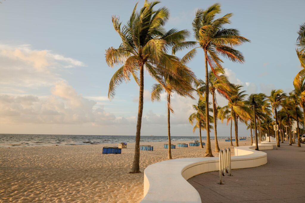 Palm trees line a sandy beach next to a curved white walkway in Fort Lauderdale, with blue trash bins and a calm ocean under a partly cloudy sky.