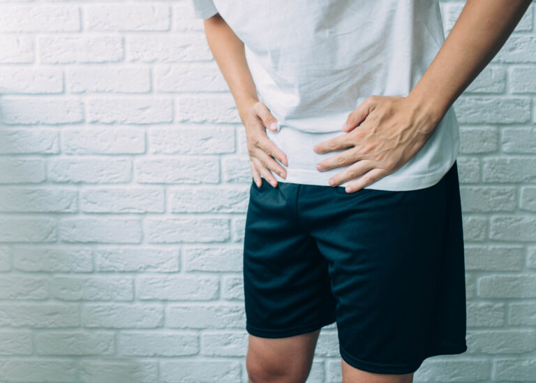 A man in a white shirt and dark shorts stands against a white brick wall, holding his lower abdomen—highlighting the need for pelvic floor therapy in Florida.