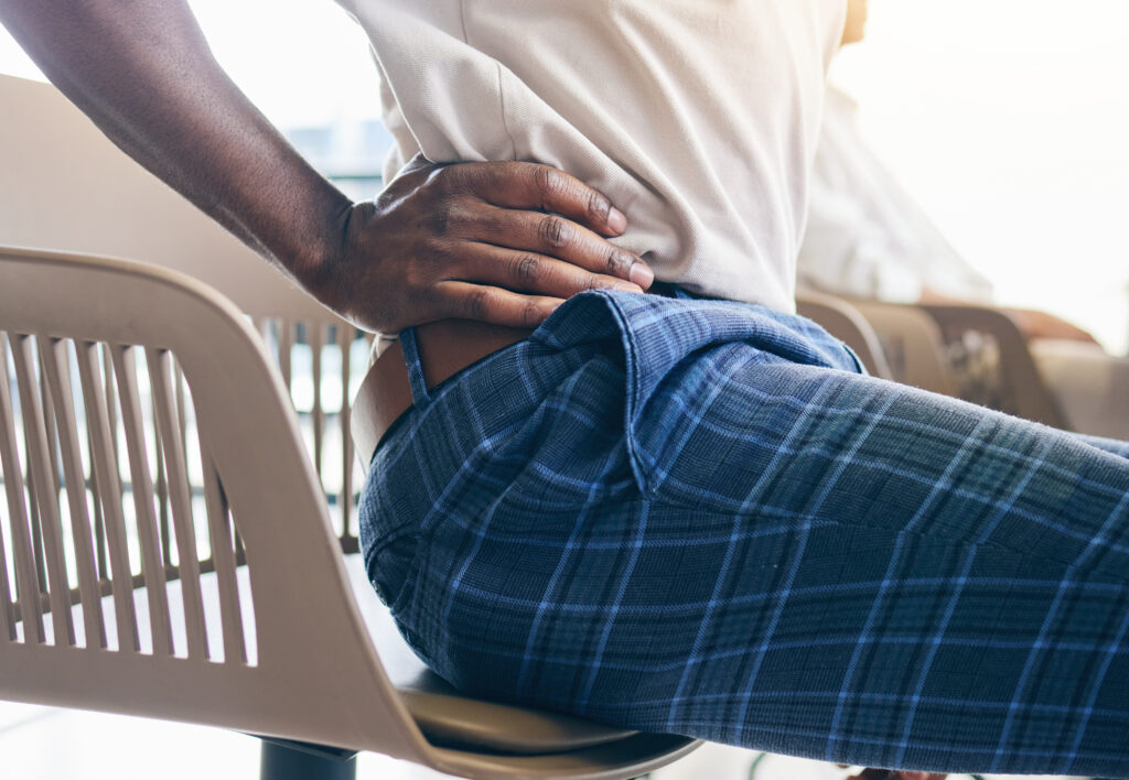 A person wearing a white shirt and plaid pants sits on a chair in Orlando, holding their lower back as if experiencing discomfort, possibly needing pelvic health or prostatitis treatment.