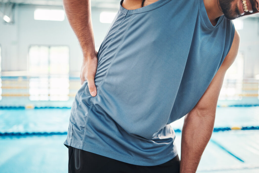 A person in athletic clothing holds their lower back, appearing to be in discomfort—possibly experiencing low back pain—inside a brightly lit gym or fitness center.