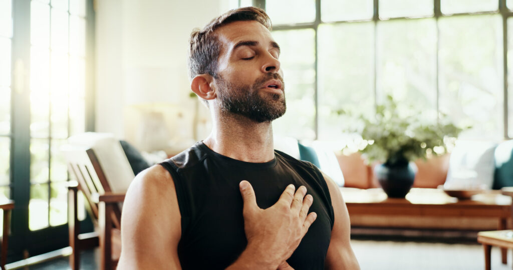 A man sits indoors with his eyes closed, wearing a sleeveless shirt and placing a hand on his chest, appearing to practice deep breathing or meditation to help manage stress and support pelvic floor health.