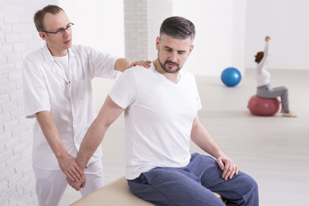 A physical therapist assists a man with arm and shoulder exercises, focusing on posture and breathing, while another person practices exercises on a stability ball in the background.