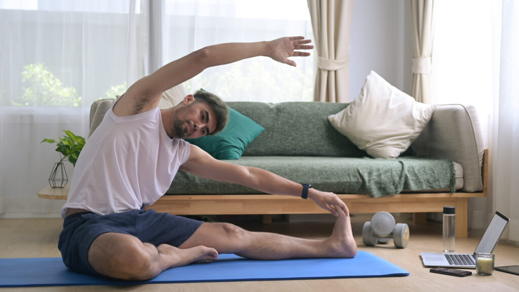 Man sitting on yoga mat in living room, stretching with one arm overhead and the other touching his foot, laptop and dumbbells nearby—an ideal setup for pain management or easing pelvic pain through mindful movement.