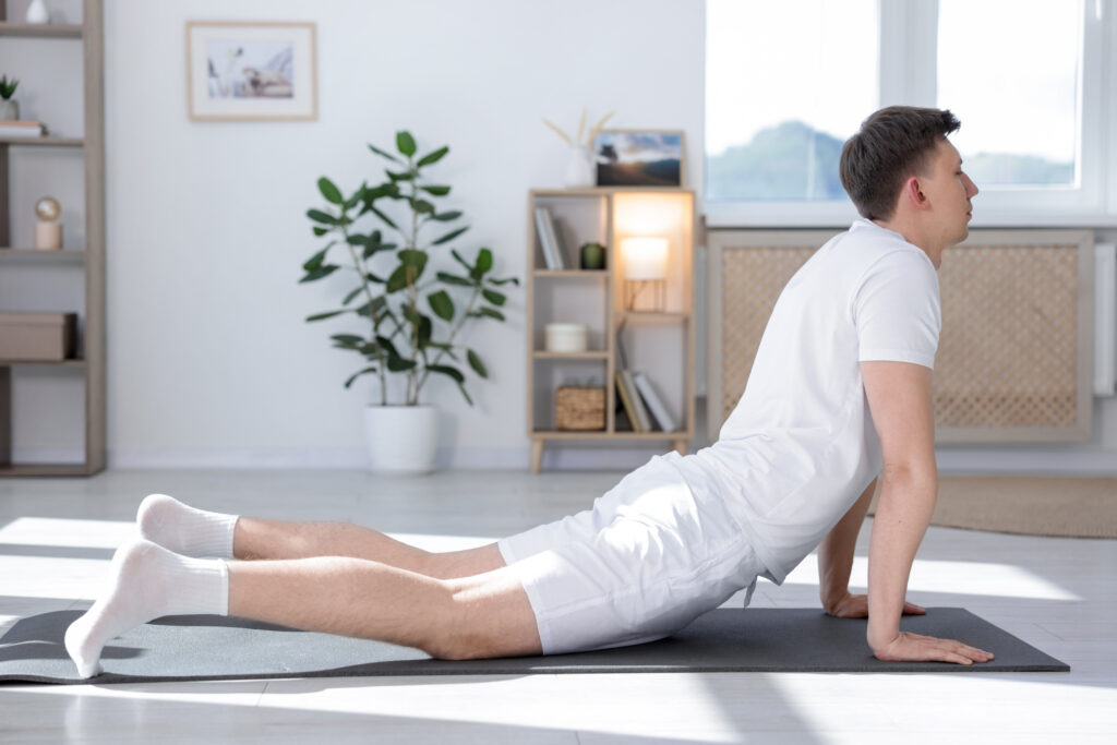 A man in white workout clothes does a cobra yoga pose on a mat in a bright living room, demonstrating an exercise often used in the treatment of pelvic floor overactivity, with plants and shelves in the background.