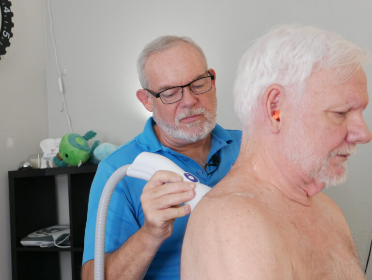 A man uses a handheld medical device on another man's upper back and shoulder in a clinical setting, demonstrating Shockwave Therapy often used to support men's health.