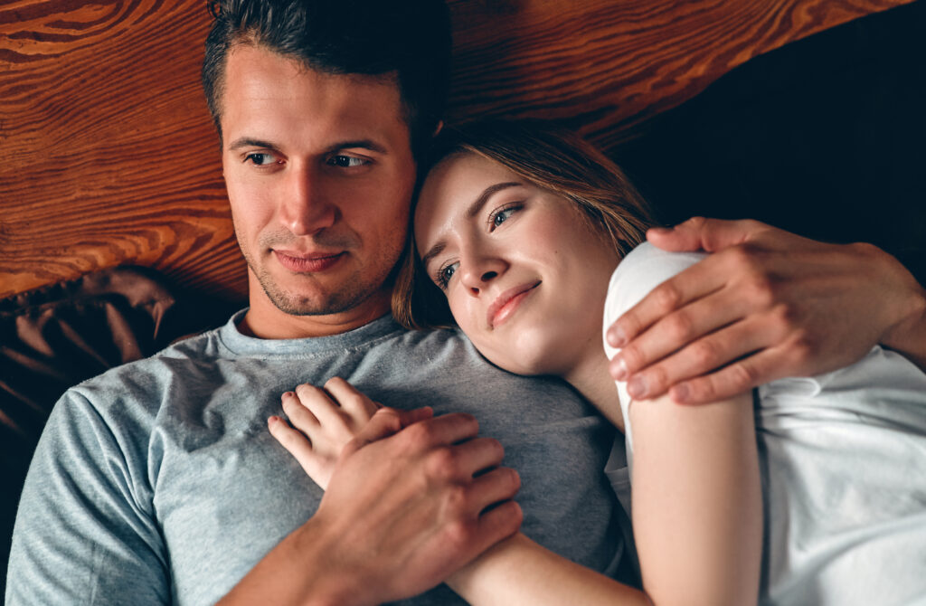 A man and a woman are lying on a bed, with the woman resting her head on the man's chest—both looking content, enjoying a quiet moment of intimacy that reflects trust and connection.