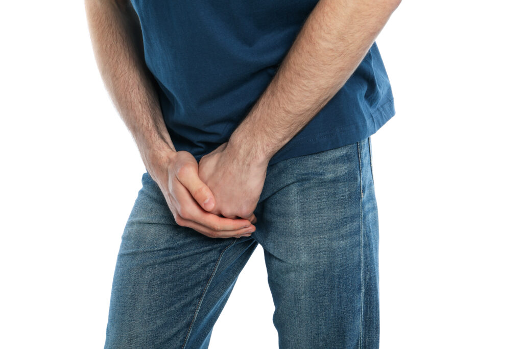 A person in a blue shirt and jeans is holding their groin area with both hands, suggesting discomfort or bladder symptoms, possibly indicating issues such as leaks.