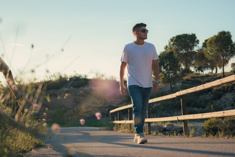 A man wearing sunglasses, a white t-shirt, and jeans walks along a paved path beside a wooden fence with trees in the background, enjoying the walking pelvic floor benefits.