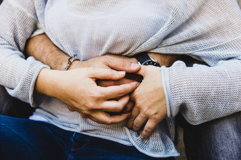 Close-up of two people sitting closely, with one person's arms wrapped around the other's torso, both wearing light-colored sweaters and blue jeans—offering comfort for those experiencing pelvic pain or pain during intimacy.