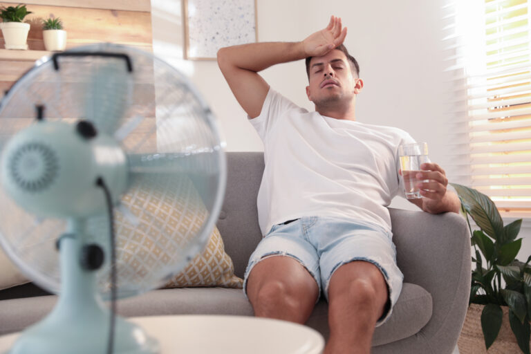 A man sits on a couch wearing a white t-shirt and shorts, holding a glass of water, and wipes his forehead in front of an electric fan as the warmer weather causes his pelvic pain to flare up.