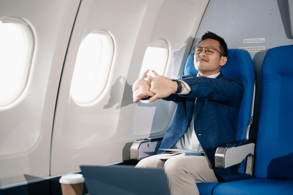 A man in business attire sits in an airplane seat by the window, stretching his arms forward with eyes closed and a laptop on the tray table—an ideal way to support pelvic health during long trips or summer travel.