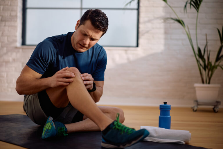 Man sitting on a mat indoors holding his knee and appearing to be in pain, with a towel and water bottle nearby—an all-too-common scene for active men and athletes seeking relief, possibly considering SoftWave Therapy.