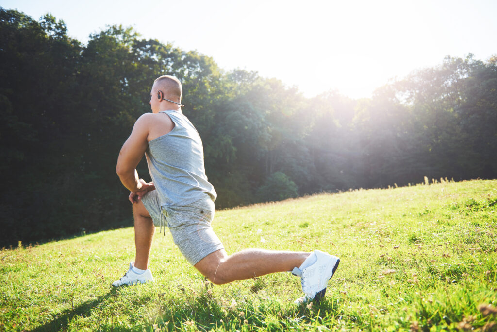 A man in athletic clothing performs a lunge stretch on a grassy field, possibly focusing on injury recovery, with trees and sunlight in the summer background.