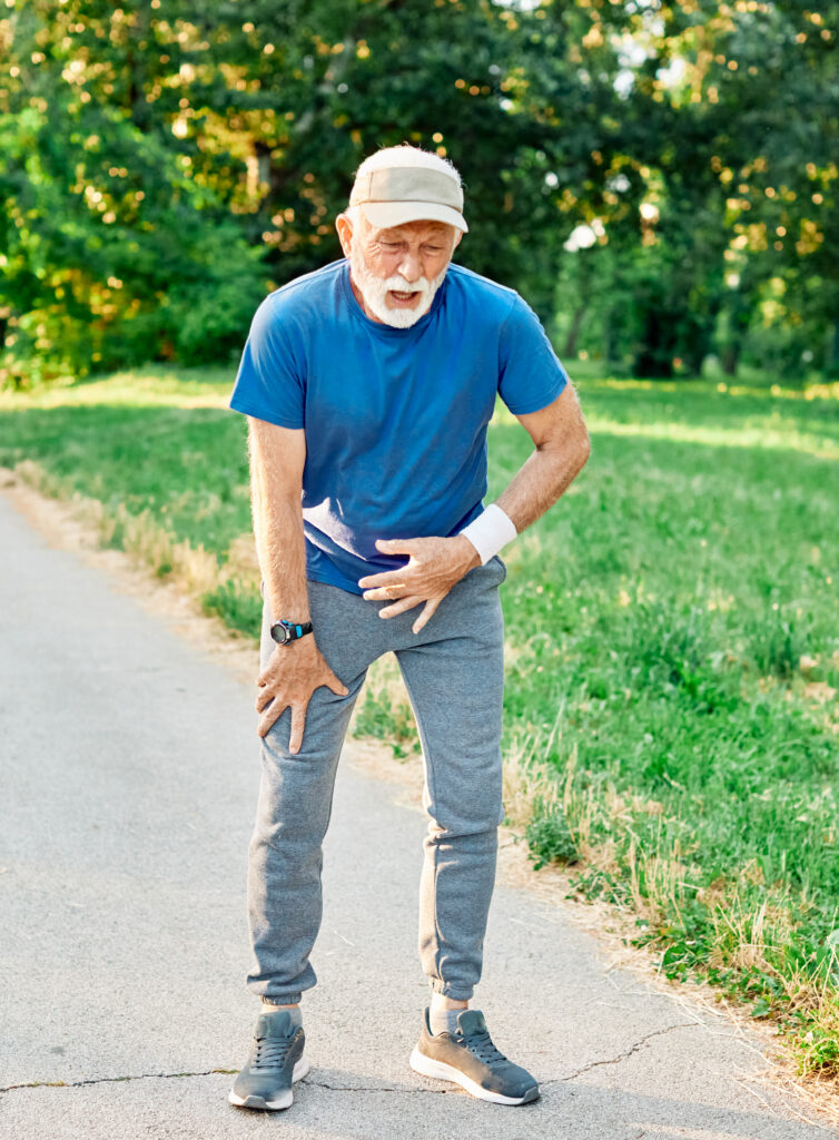 An older man in athletic clothes stands on a path outdoors, gripping his knee with a pained expression—struggling with discomfort, possibly worsened by the Florida heat and humidity.