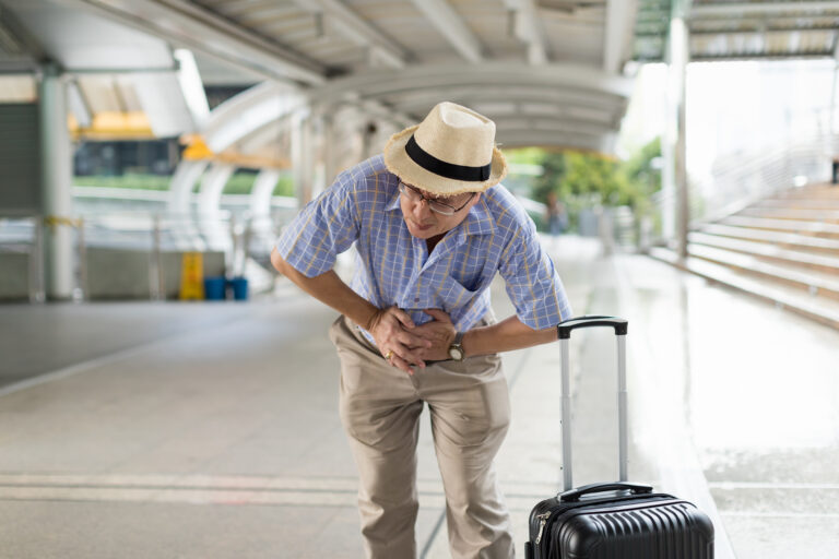 An older man wearing a hat and glasses clutches his stomach in pain, possibly experiencing pelvic pain, while standing next to a suitcase at an outdoor transit terminal.