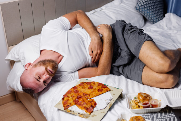 A man lies on a bed holding his stomach in discomfort, likely experiencing constipation after indulging in summer eating, with an open pizza box and other fast food containers beside him.