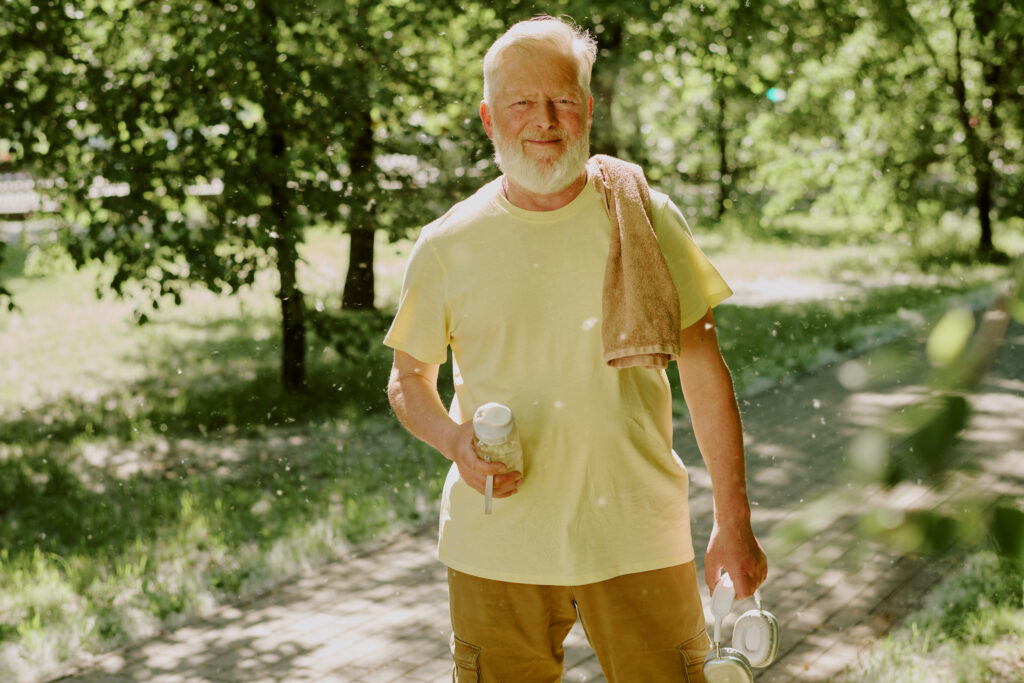 Older man in a yellow shirt stands outdoors on a sunny day, staying cool with a water bottle for hydration and a portable fan. A towel rests over his shoulder, as he enjoys the sunlight while caring for his bladder health. Trees and sunlight fill the background.