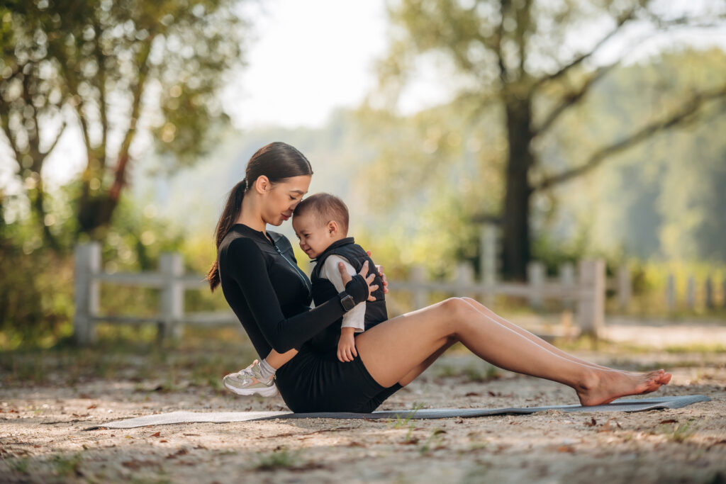 A woman in athletic wear sits on an exercise mat outdoors, holding a small child in her lap as they touch foreheads and smile—celebrating safe summer movement perfect for postpartum moms.