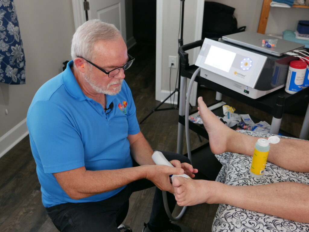 A man in a blue shirt uses a handheld device to perform shockwave therapy on a person's foot in a clinical setting. Medical equipment and supplies are visible on a nearby cart, highlighting the blend of modern techniques with traditional treatments.