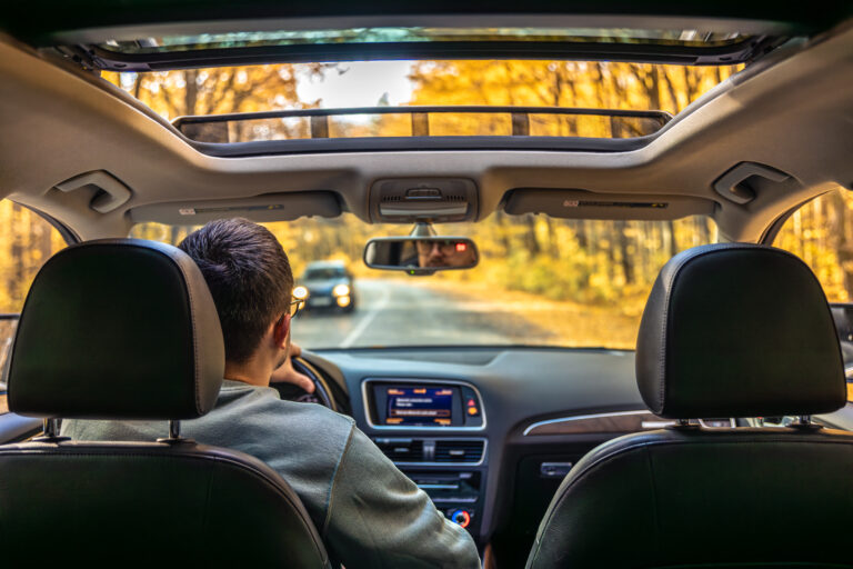 View from the backseat of a car with a sunroof, showing a person driving on a road lined with autumn trees and another car approaching ahead—an inviting reminder that sitting too much during long drives, whether in autumn or on summer road trips, calls for mindful movement to support your pelvic floor health.