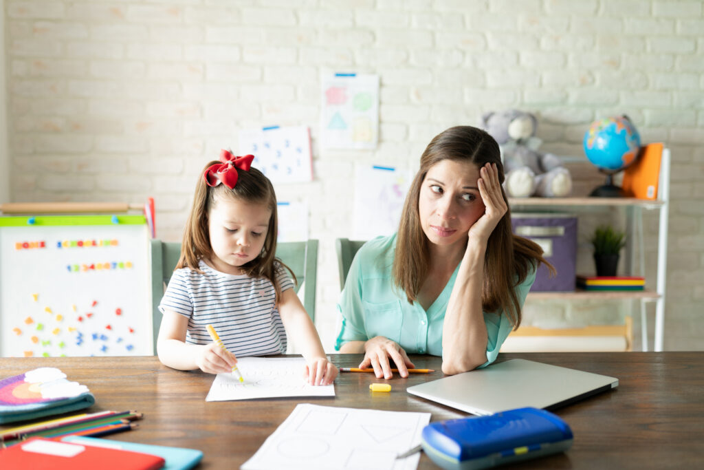 A young girl draws while an adult woman, perhaps feeling back-to-school stress, sits beside her—looking tired and resting her head on her hand at a cluttered table with school supplies.