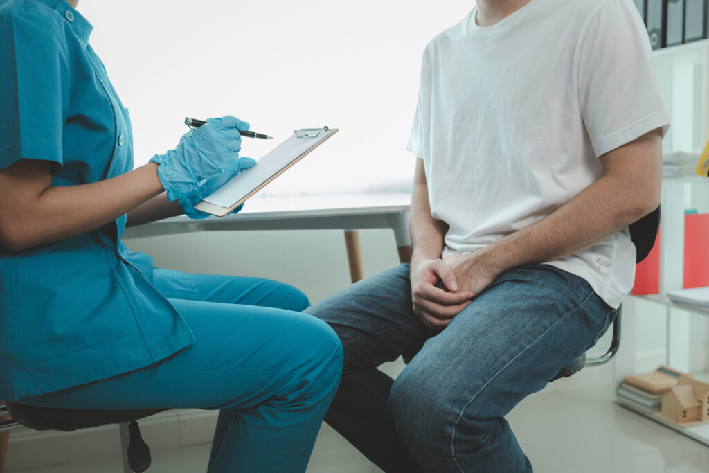 A healthcare worker in blue scrubs holds a clipboard while sitting across from a person in a white shirt and jeans in a medical office, discussing important pelvic health signs as part of a men’s health consultation.