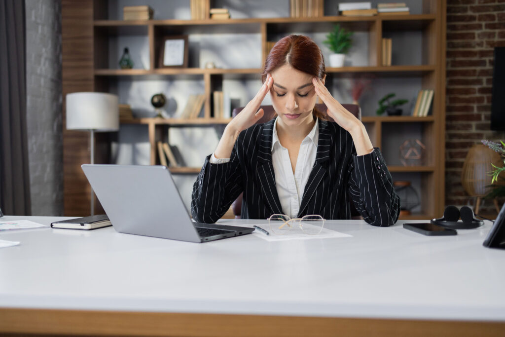 A woman in a suit sits at a desk with a laptop, holding her head in her hands and looking stressed. Papers and glasses are on the desk, and a bookshelf is in the background—reflecting how chronic stress can impact the nervous system and even contribute to symptoms like pelvic pain.