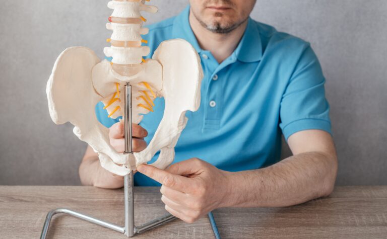 A man in a blue shirt sits at a table, holding and pointing to a human pelvic bone model with a metal stand, showing the lower spine and pelvis—an area often discussed in connection with urinary leakage in men or issues such as post-void dribble.